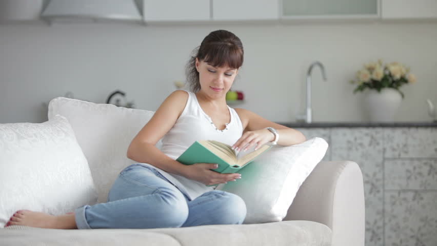Pretty young woman relaxing on sofa reading book and smiling at camera