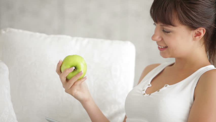 Young woman sitting on sofa with touchpad eating apple and smiling at camera