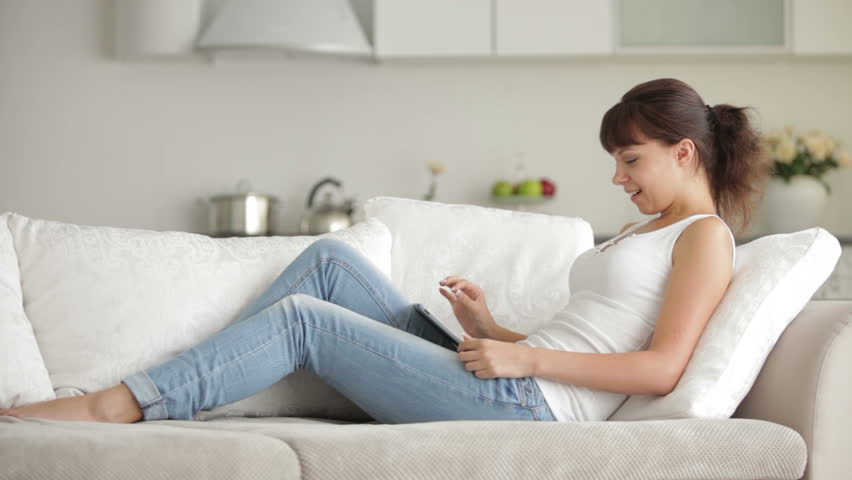 Cute girl sitting on sofa with touchpad and credit card and smiling at camera