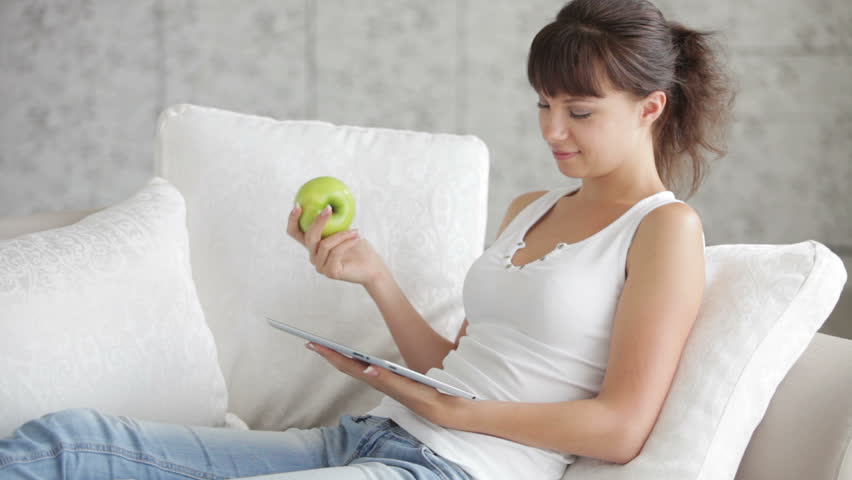 Cute girl relaxing on sofa using touchpad and eating apple
