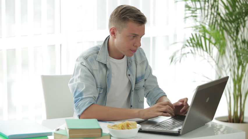 Handsome student sitting at table using laptop and eating chips