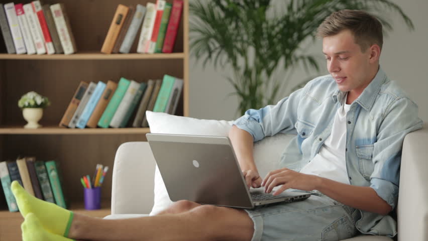 Handsome young student sitting on sofa and using laptop
