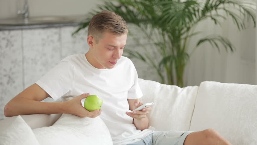 Handsome young men sitting on sofa eating apple and using cellphone