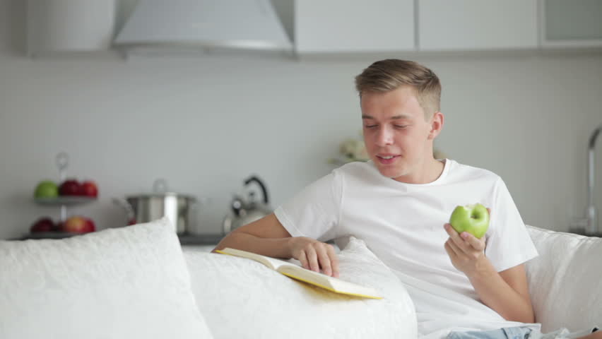 Handsome young men sitting on sofa reading book and eating apple