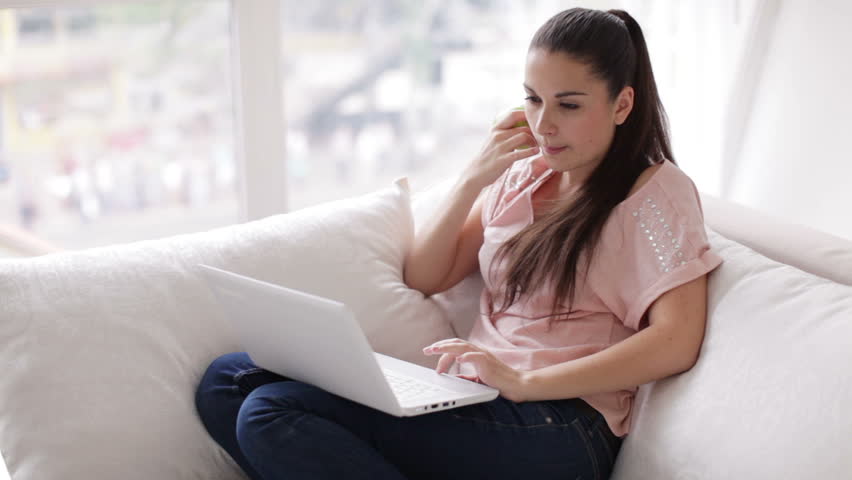 Beautiful girl sitting on sofa using laptop eating apple and smiling