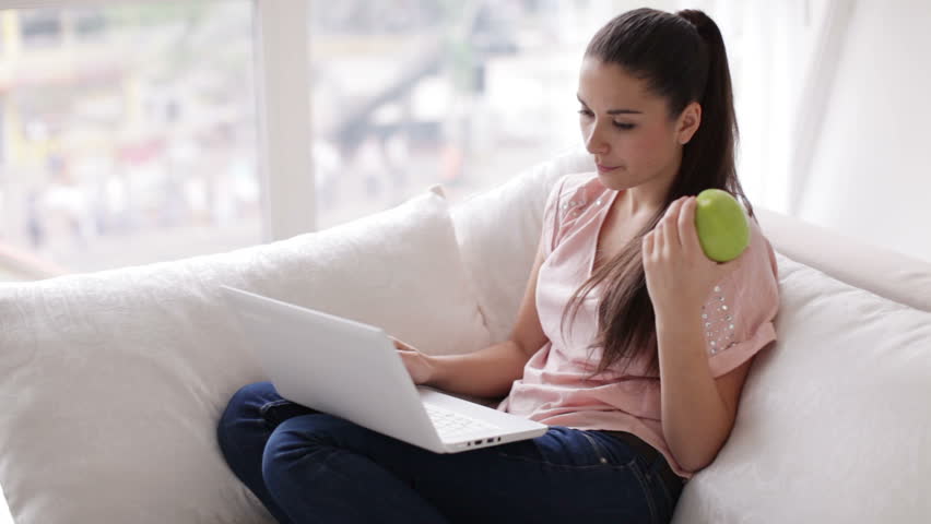 Beautiful girl sitting on sofa with laptop holding apple and smiling