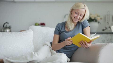 Beautiful young woman sitting on sofa reading book and smiling