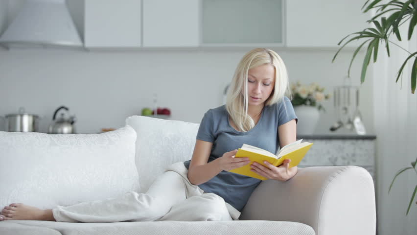 Charming young woman sitting on sofa with book and smiling at camera