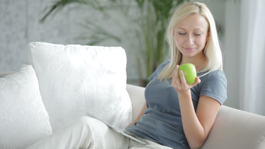 Charming young woman sitting on sofa using touchpad holding apple and smiling