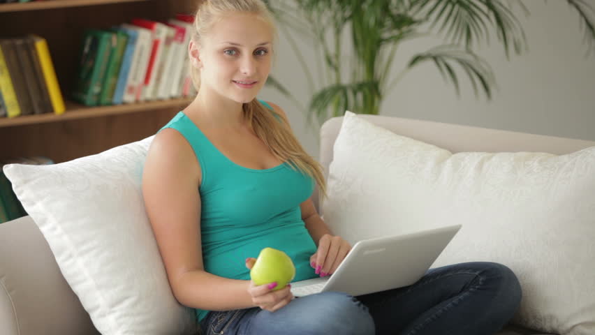 Cute girl sitting on sofa with laptop and eating apple