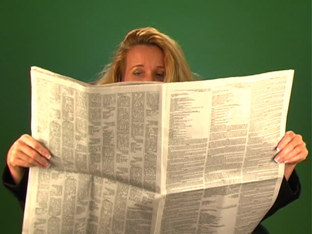 A lovely blonde businesswoman, sitting in an office chair with a plain background, reads a newspaper.