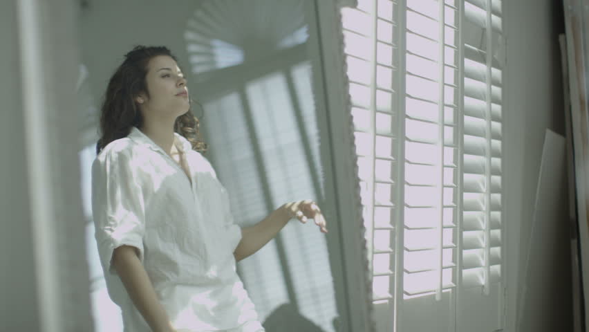 Beautiful young woman relaxing in her elegant apartment. Sunlight streams in as she opens the window shutters and she sits down to look at the view outside. In slow motion.