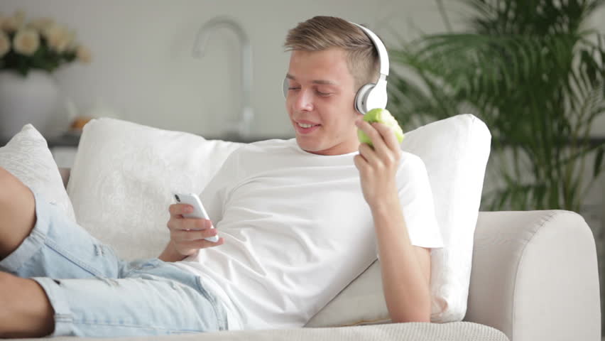 Beautiful guy relaxing on sofa using cellphone listenning music and eating apple