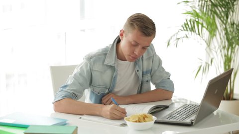 Attractive student sitting at table using laptop and eating chips