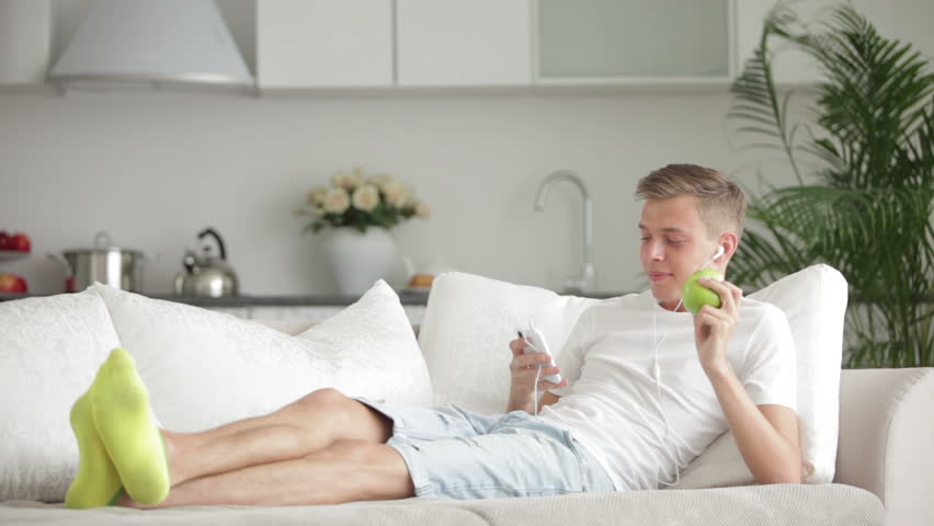 Attractive young man lying on sofa using cell phone and eating apple