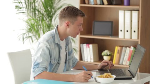 Attractive man student sitting at table using laptop and eating chips