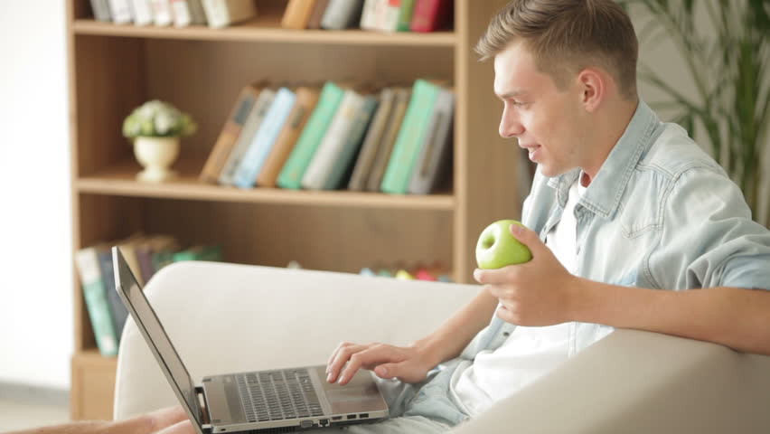 Handsome man sitting on sofa using laptop and eating apple