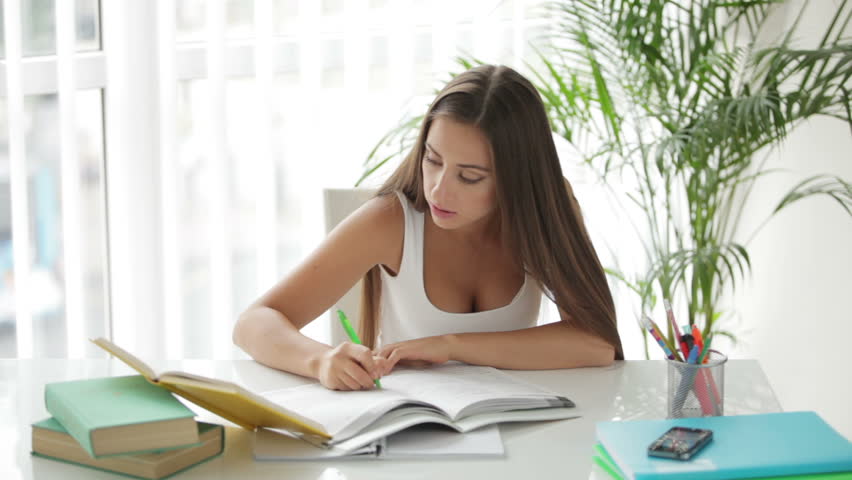 Young girl sitting at table reading book and writing in notebook