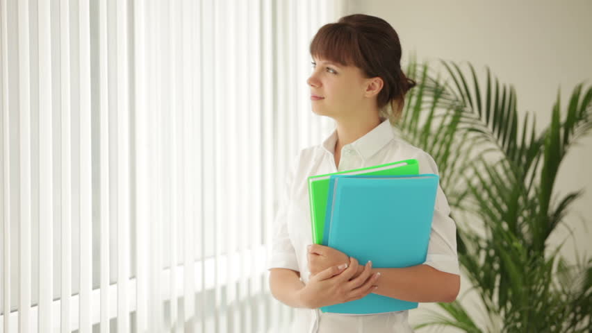 Attractive business woman standing in front of window holding paper folders