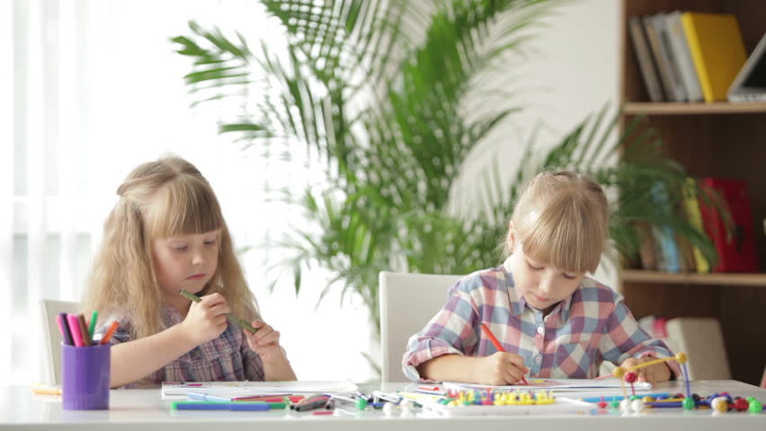 Two cute little girls sitting at desk drawing with colored pencils and smiling