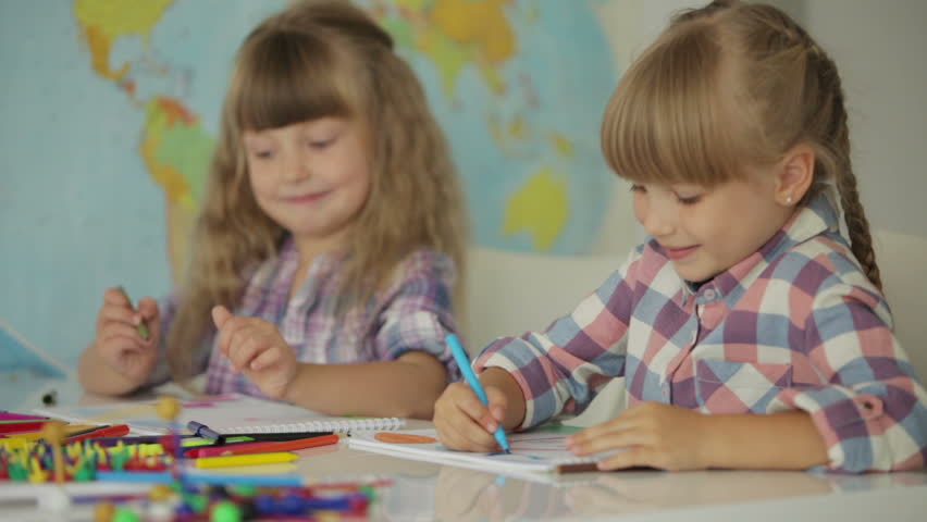 Two funny little girls sitting at desk drawing with colored pencils