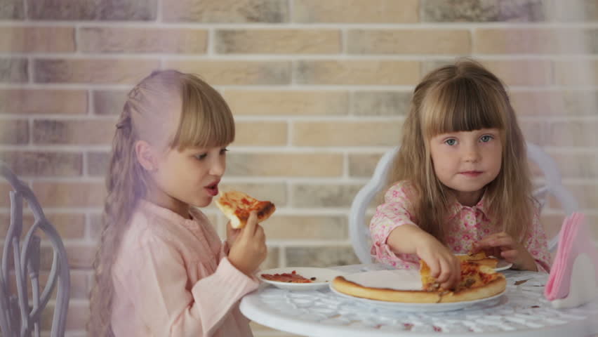 Two cute little girls sitting at table at cafe eating pizza and smiling