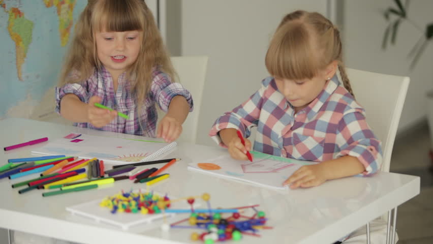 Two cute little girls sitting at table drawing with colored pencils and smiling