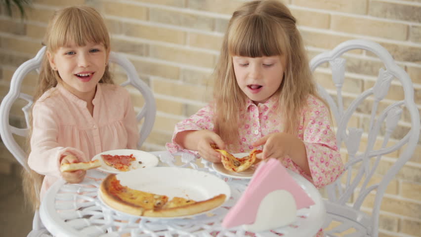 Two funny little girls sitting at table at cafe eating pizza and smiling