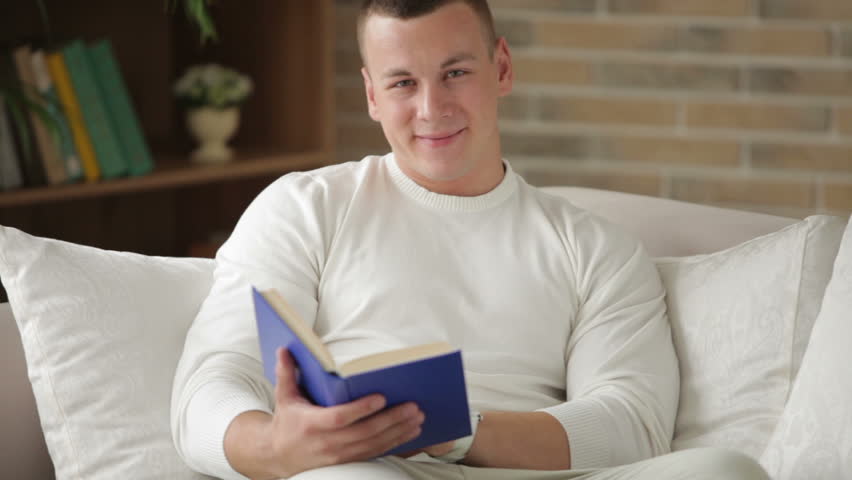 Good-looking guy sitting on couch reading book and smiling