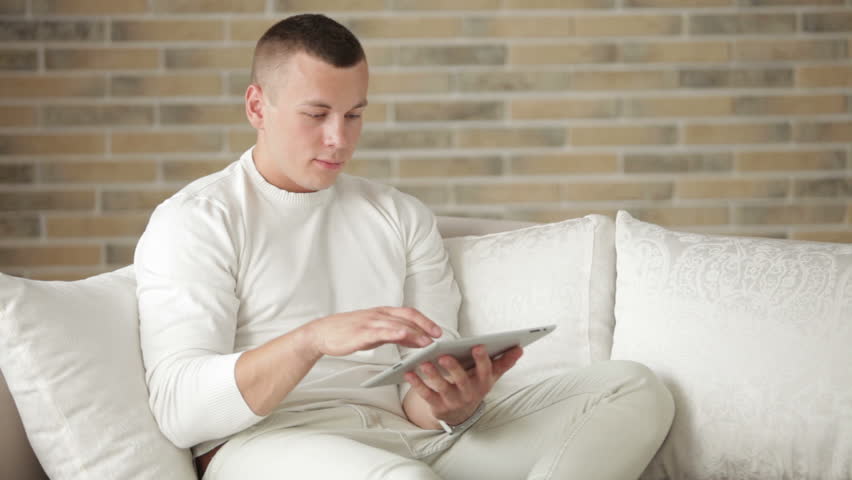 Good-looking guy relaxing on sofa using touchpad and smiling at camera