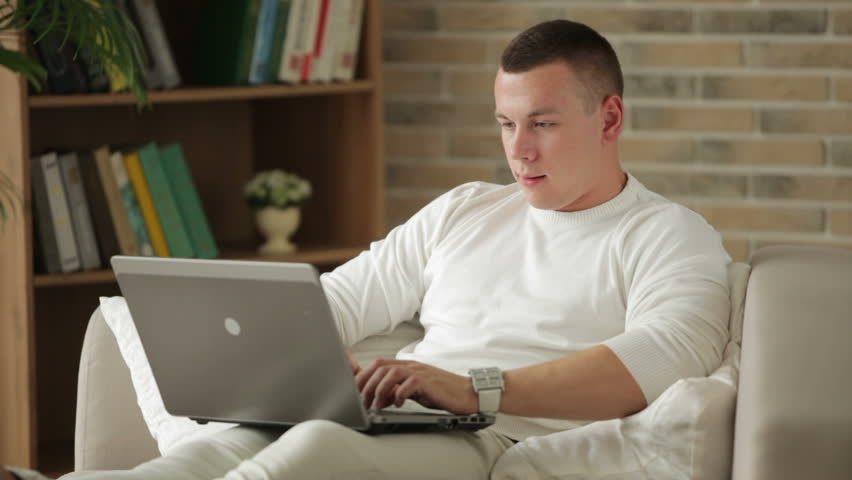 Good-looking guy sitting on sofa using laptop and smiling