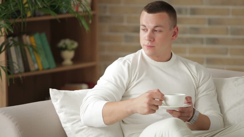 Good-looking guy sitting on sofa holding cup of tea and smiling