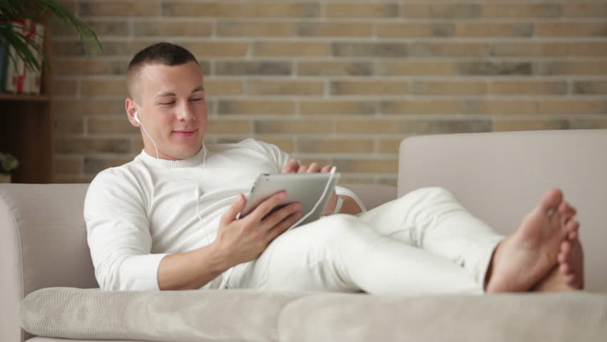 Handsome guy in headset lying on sofa using touchpad and smiling