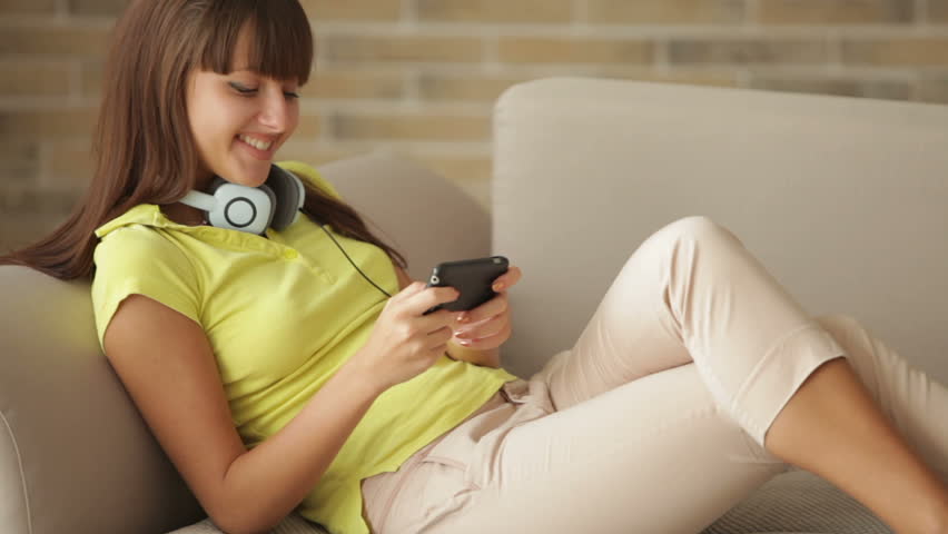 Cute girl sitting on sofa using cellphone and smiling at camera