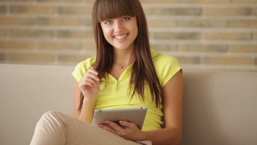 Cute girl sitting on couch using touchpad and smiling at camera