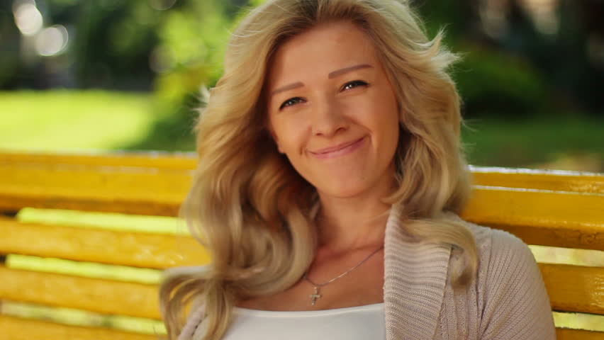 Pretty blond female smiles in park on bench, looks at camera