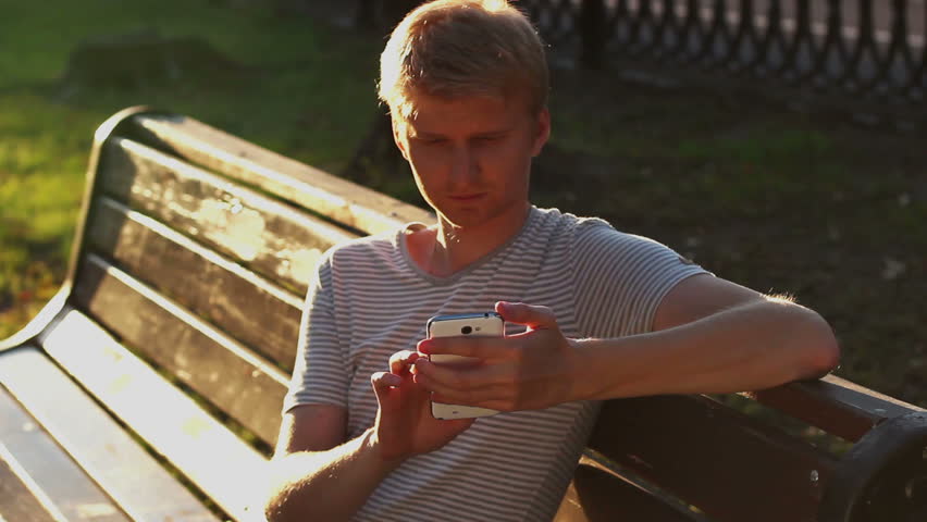Young blond adult man writing sms message in park bench sitting