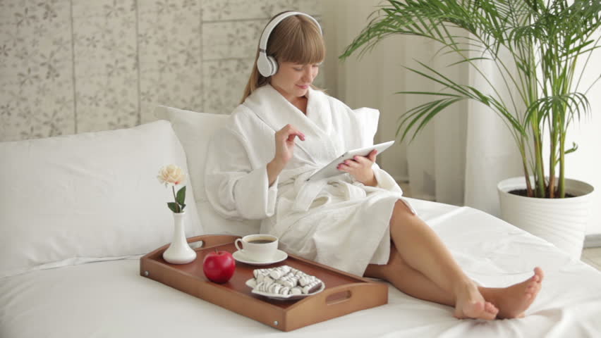 Cheerful young woman in headset relaxing on bed and using touchpad
