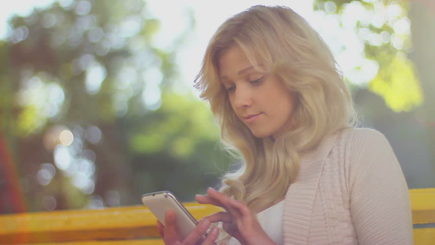Woman types sms smiles sending message, daytime park bench