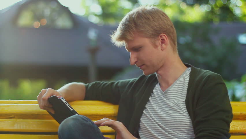 Young blond male reading tablet pc electronic book in park bench