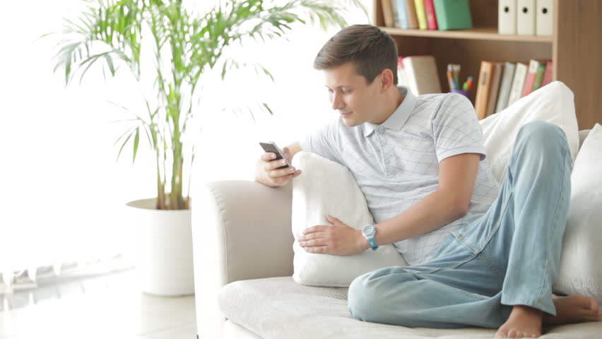Good-looking guy sitting on sofa using mobile phone looking at camera and