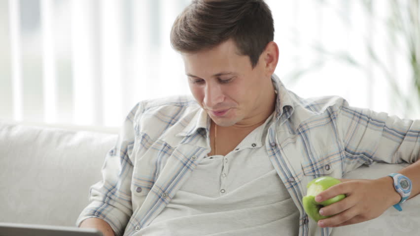 Attractive guy sitting on sofa eating apple using laptop and smiling at camera