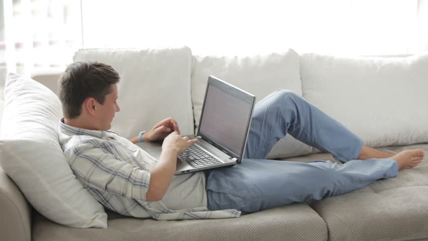Attractive guy relaxing on sofa using laptop closing it and looking at camera