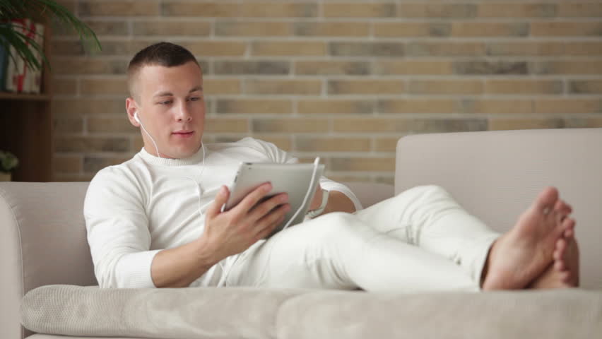 Young man in earphones lying on sofa and using touchpad