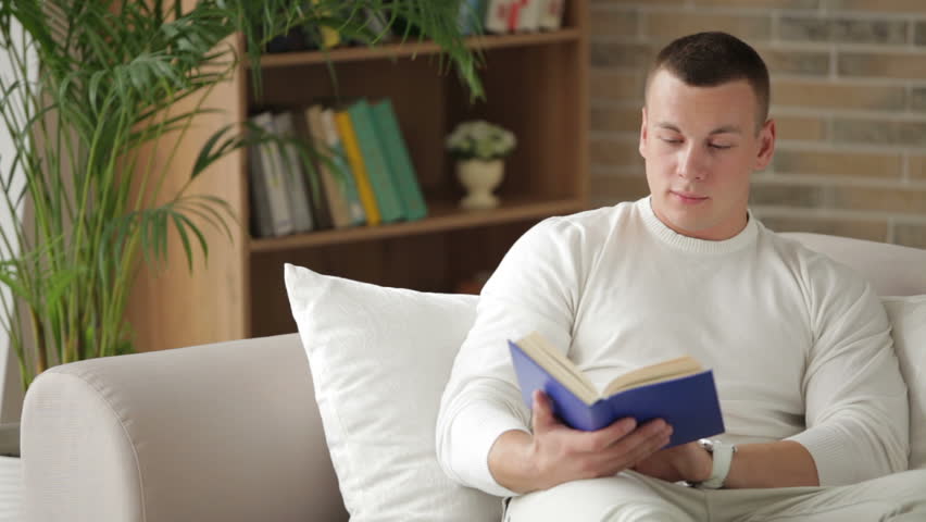 Young man sitting on couch with book and looking at camera with smile