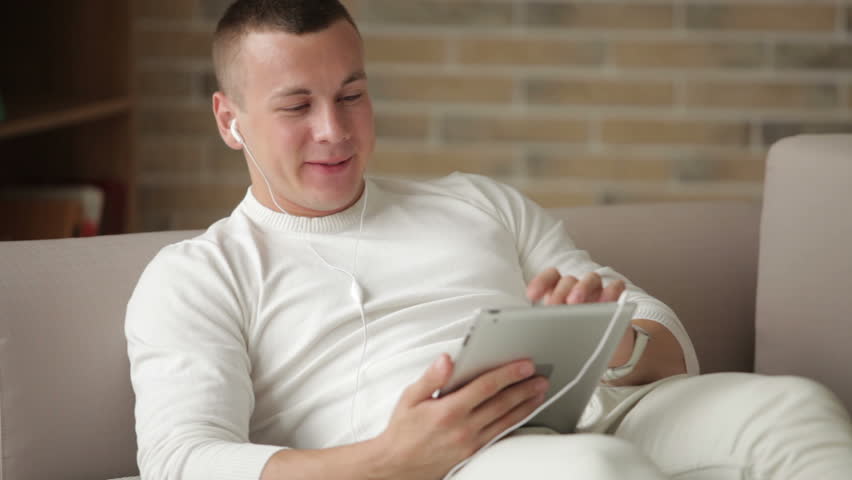 Smiling guy in headphones lying on sofa and using touchpad
