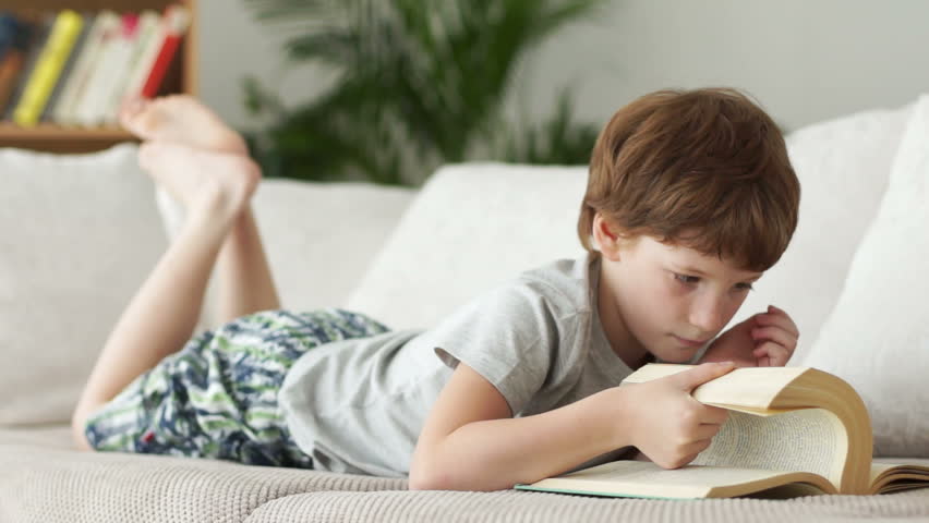 Funny little boy lying on sofa and turning pages of book