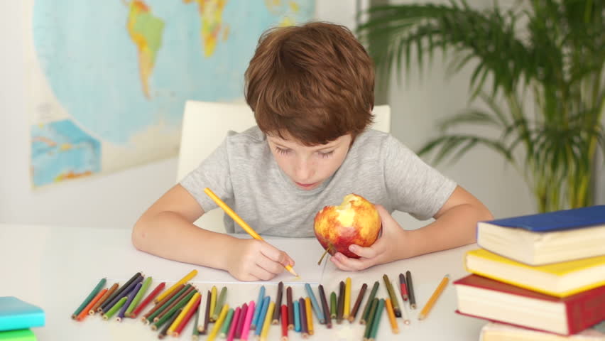 Little boy sitting at table holding apple and drawing with colored pencils