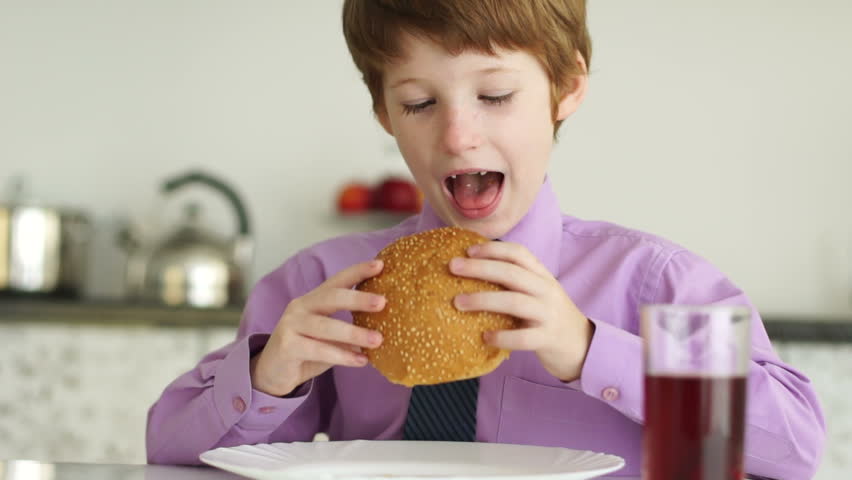Funny little boy sitting at table and eating burger