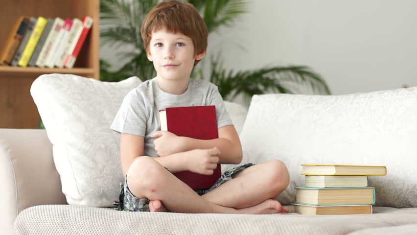 Cute little boy sitting on sofa holding book and showing thumb up